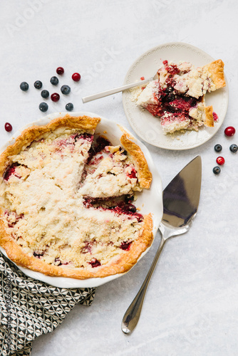 A berry pie with two slices taken out. One slice is still in the pie pan and the other is on a plate to the side. There is a pie serving utensil and berries all around. A delicious treat.