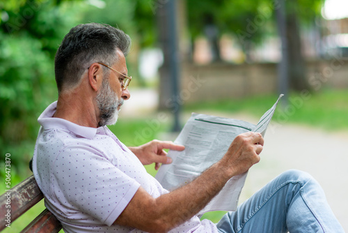 Side view of mature man seated on a wooden bench reading a newspaper in a park. Senior man reading newspaper in the park. Senior gray-haired man in glasses.
