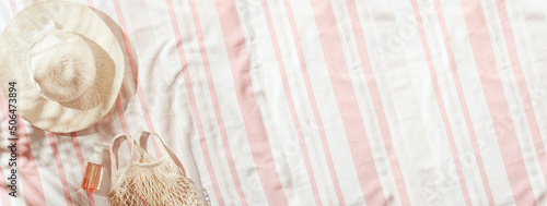 Fotografia Summer minimal wide banner, top view wide-brimmed sun hat, cosmetics products on striped beach towel, pale pink background, sun shadows from palm leaf