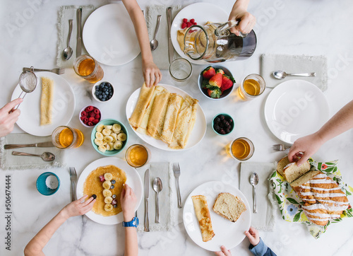 A family of 6 is sitting around a table serving each other breakfast. There are pre-made crepes in the middle of the table. There is fruit all around such as strawberries, raspberries, blueberries, bl