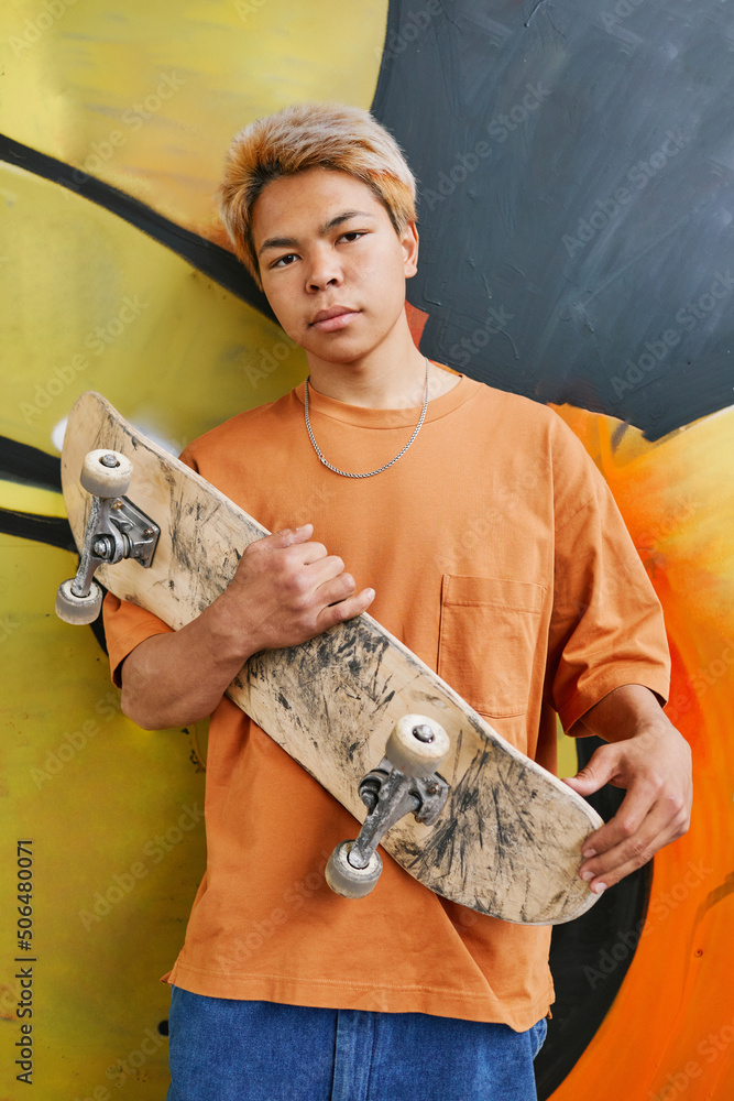 Colorful portrait of young teenage boy with skateboard standing against graffiti wall in orange and looking at camera - obrazy, fototapety, plakaty 