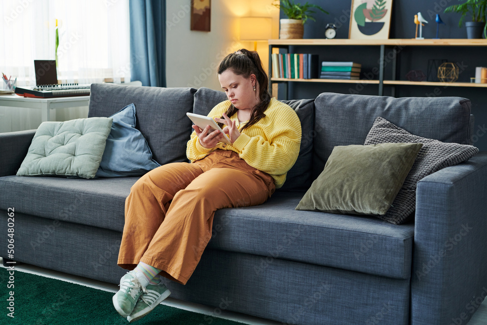 Obraz premium Portrait of modern girl with Down syndrome sitting on sofa in living room at home using digital tablet