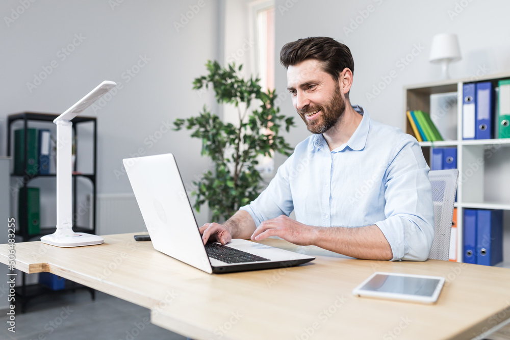 Portrait of successful businessman, man smiling and working on laptop in modern bright office
