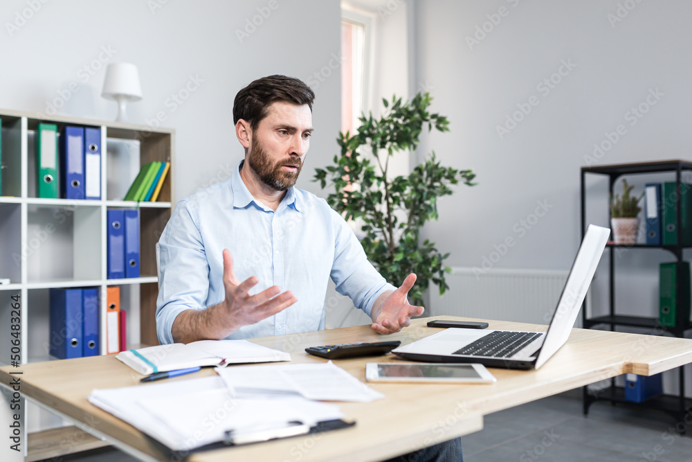 Annoyed, emotional, nervous young man, office worker, manager talking on a video call at the computer, sitting at a table in the office in white clothes Annoyed, emotional, nervous young man, office worker, manager talking on a video call at the computer, sitting at a table in the office in white clothes