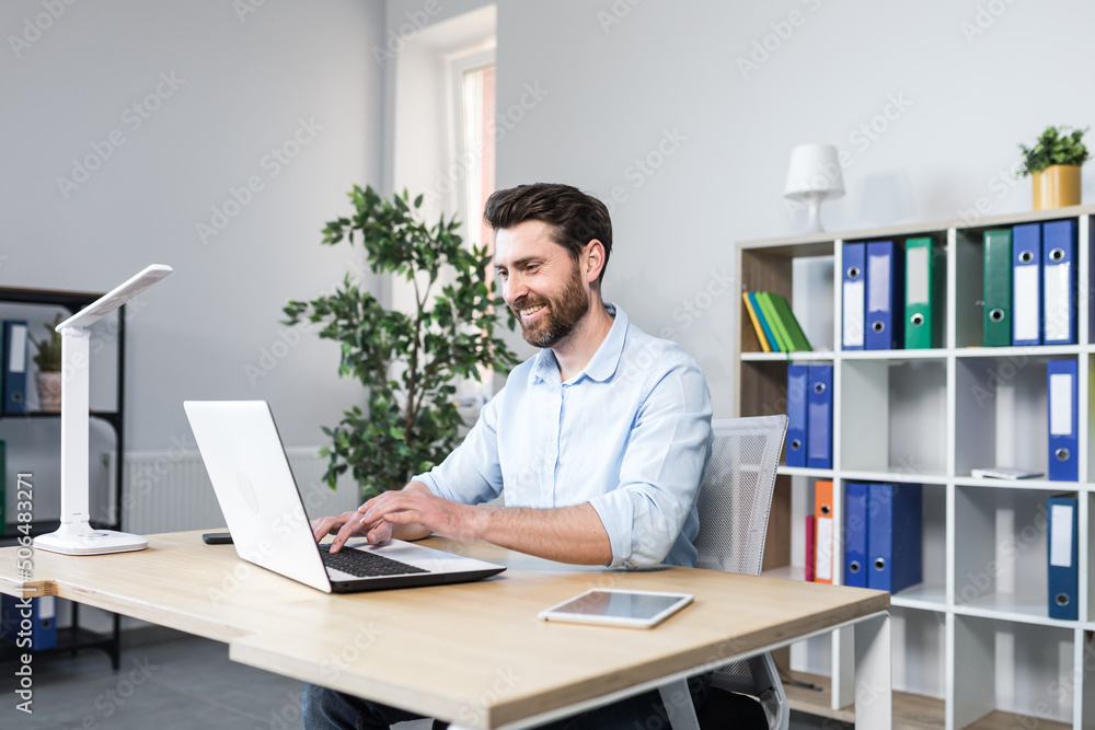 Portrait of successful businessman, man smiling and working on laptop in modern bright office Portrait of successful businessman, man smiling and working on laptop in modern bright office