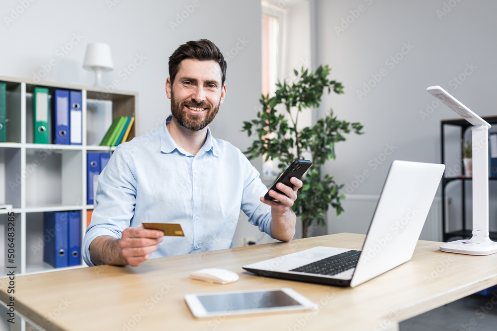 Happy businessman makes online purchases working in the office, husband holds credit bank card and uses laptop Happy businessman makes online purchases working in the office, husband holds credit bank card and uses laptop