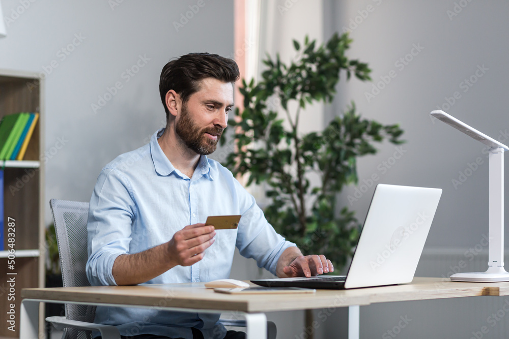 Happy businessman makes online purchases working in the office, husband holds credit bank card and uses laptop Happy businessman makes online purchases working in the office, husband holds credit bank card and uses laptop