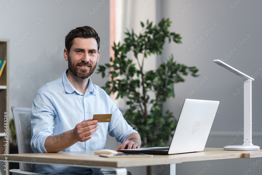 Happy businessman makes online purchases working in the office, husband holds credit bank card and uses laptop Happy businessman makes online purchases working in the office, husband holds credit bank card and uses laptop