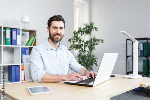 Happy businessman working in bright modern office behind laptop man with beard smiling and looking at cameras