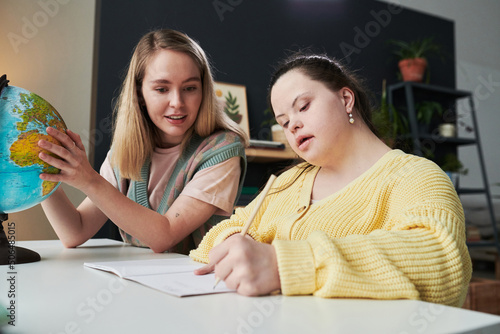 Young female teacher sitting at table next to her student with Down syndrome helping with task during geography lesson photo