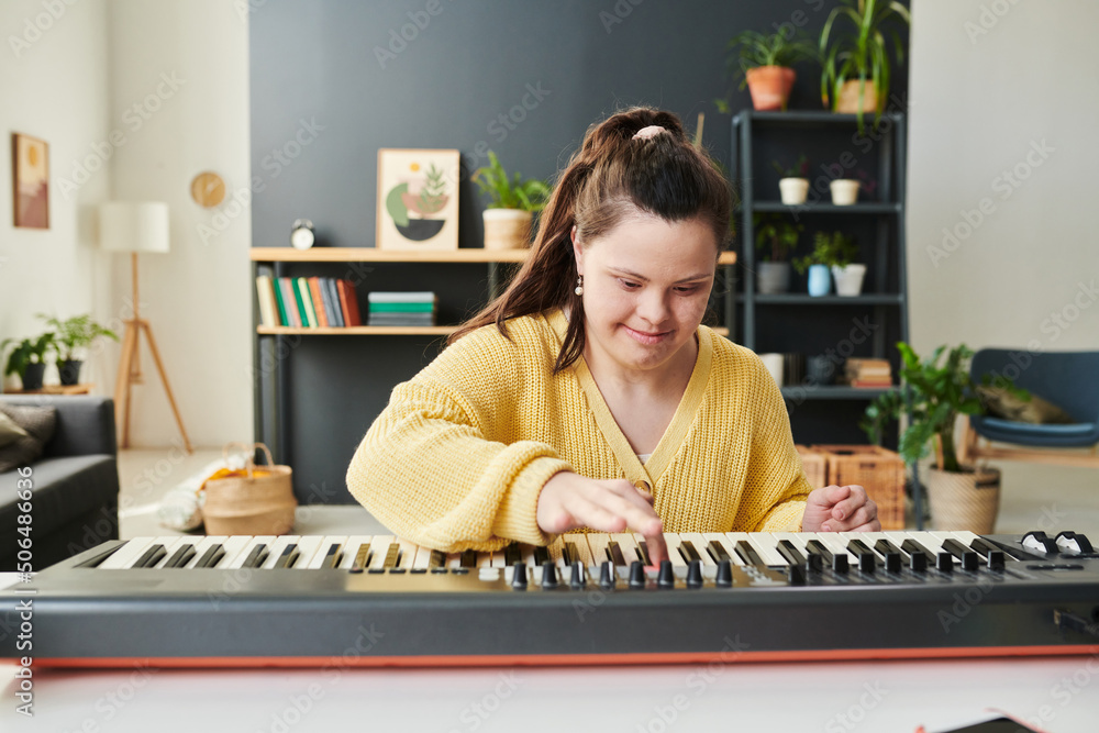 Portrait of joyful young Caucasian girl with Down syndrome spending time at home learning to play synthesizer