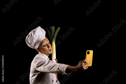Young female comedian in chef outfit taking selfie with leek