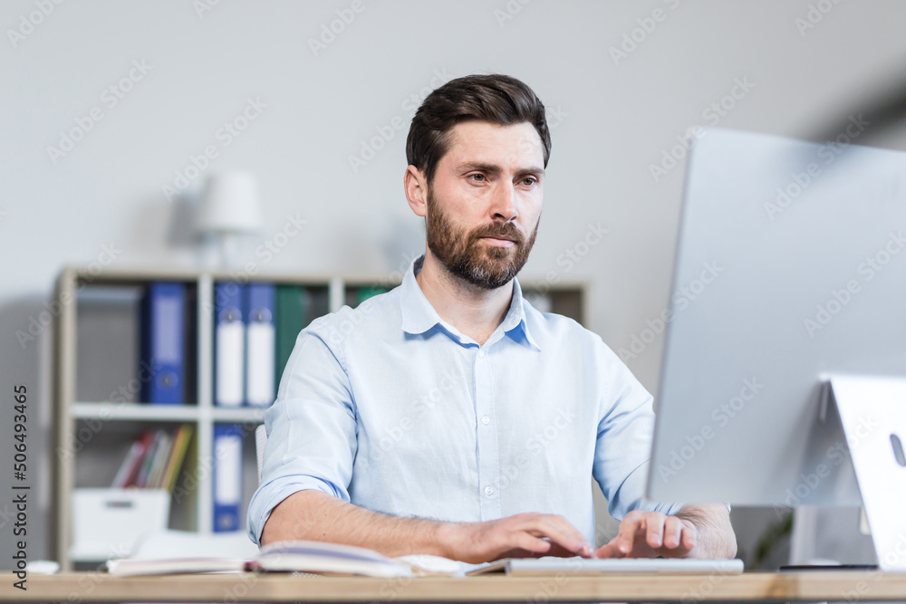 Portrait of a young man, focused and serious office worker, manager, director working at a computer, sitting behind a desk in the office Portrait of a young man, focused and serious office worker, manager, director working at a computer, sitting behind a desk in the office