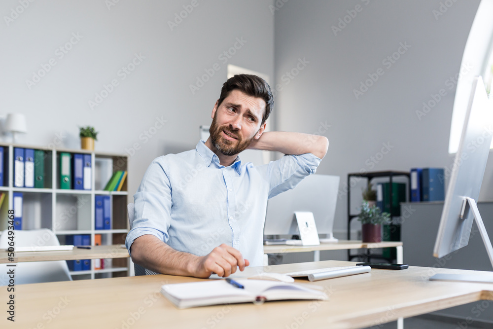 A tired young man working at a computer and sitting at a desk in the office feels a pain in the neck, massages his neck, relaxes. A tired young man working at a computer and sitting at a desk in the office feels a pain in the neck, massages his neck, relaxes.