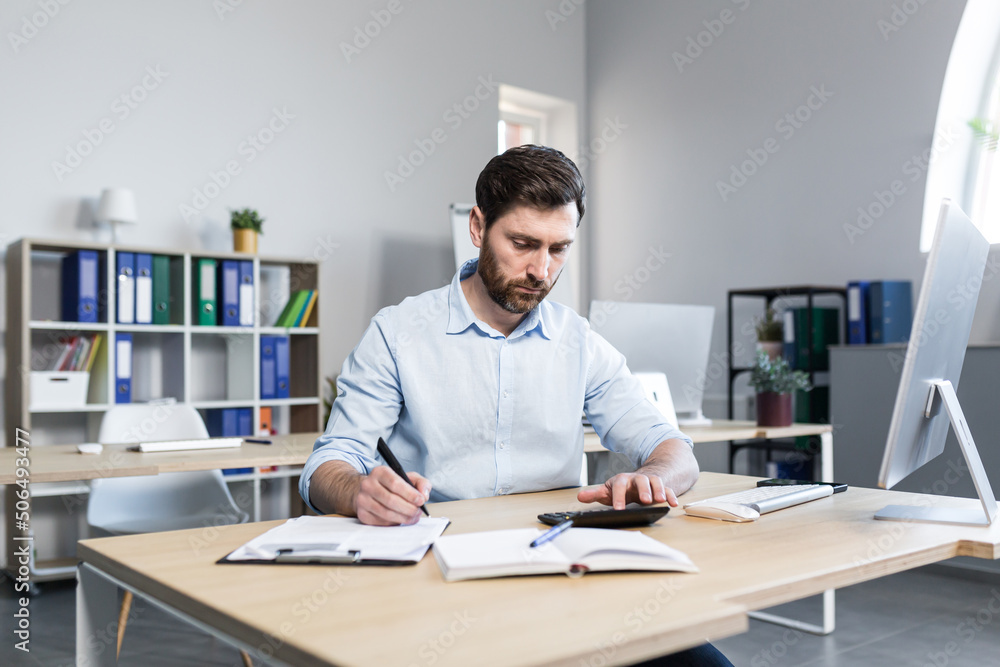 Portrait of a young man. Accountant, economist, financier. Counts on a calculator, sits at a desk at the computer, works with documents Portrait of a young man. Accountant, economist, financier. Counts on a calculator, sits at a desk at the computer, works with documents