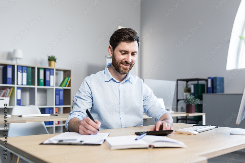 Portrait of a young man. Accountant, economist, financier. Counts on a calculator, sits at a desk at the computer, works with documents Portrait of a young man. Accountant, economist, financier. Counts on a calculator, sits at a desk at the computer, works with documents