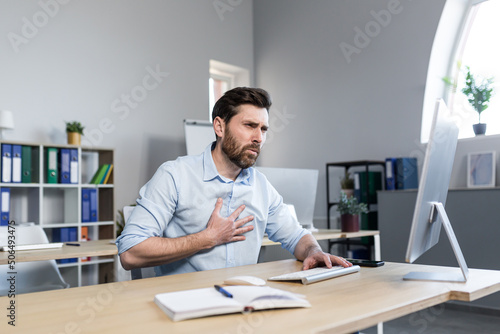 A tired young man working at a computer and sitting at a desk in the office, feels chest pain, shortness of breath, difficulty breathing photo