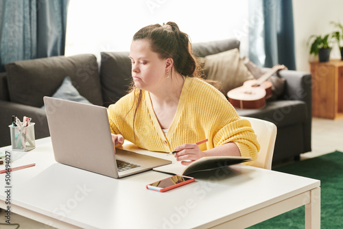 Portrait of young female university student with Down syndrome sitting at table at home using laptop to do homework photo