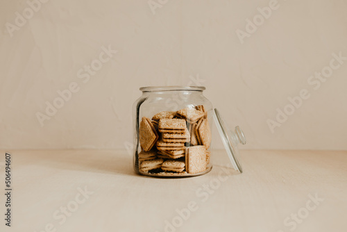 A glass jar of tasty biscuit on wooden table with beige background. Wholemeal cookies for afternoon tea. Copy space. The lid is off the jar, ready to eat the cookies. 