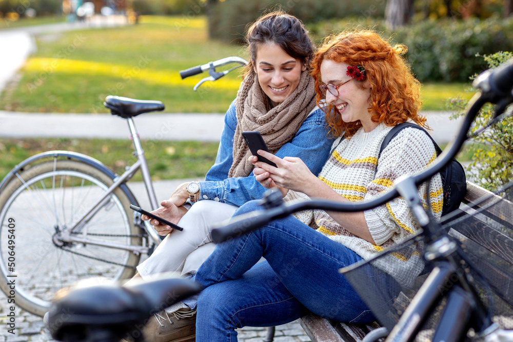 Two friends woman sitting on the bench and looking at mobile phone and smile