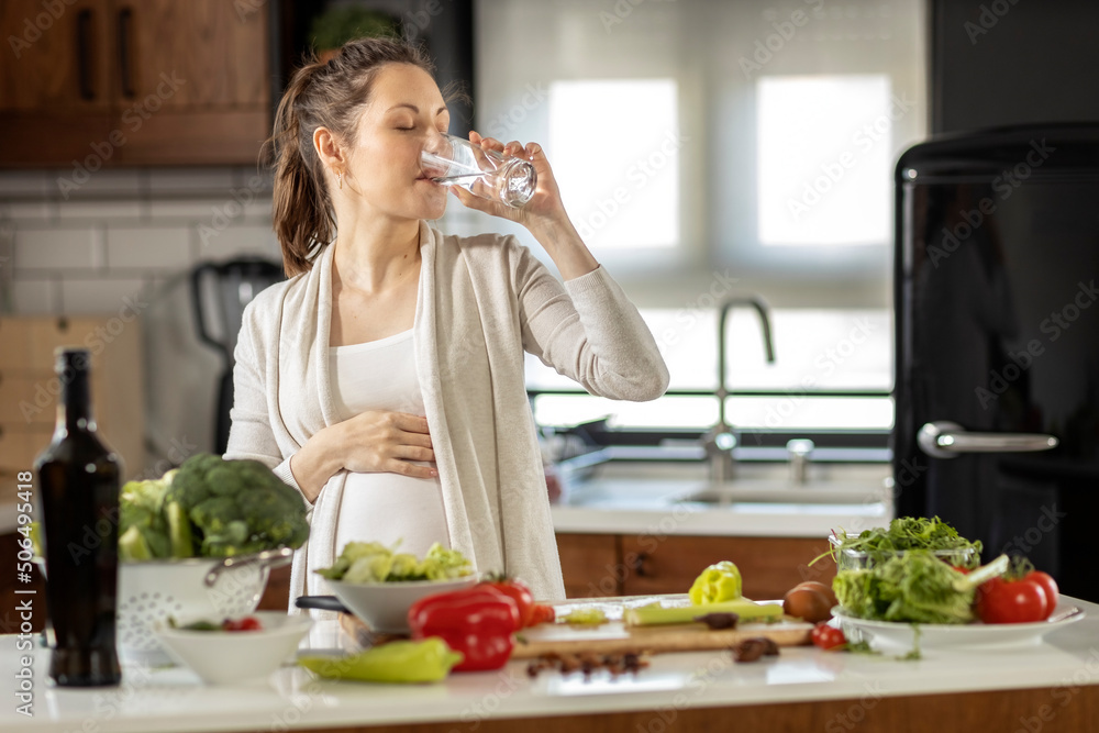 Pregnant woman in the kitchen prepare vegetarian healthy meal and drink water from glass