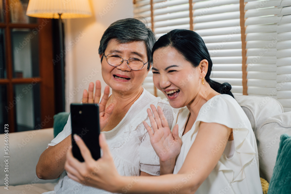 Merry Asian senior mom and adult daughter smiling and waving hands while sitting on sofa and making video call via cellphone in daytime at home Merry Asian senior mom and adult daughter smiling and waving hands while sitting on sofa and making video call via cellphone in daytime at home