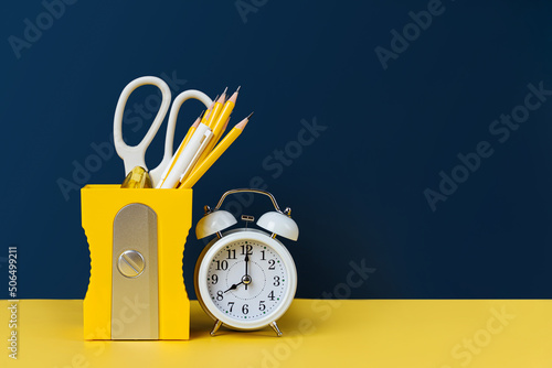 Obraz na plátně Yellow pencil holder and alarm with school stationery supplies on blue background