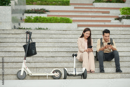 Smiling young businesss couple sitting outdoors next to e-scooter and checking social media on smartphones photo