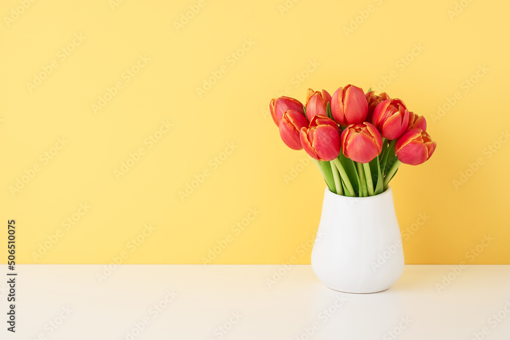 Bunch of red tulips in a white ceramic vase on the white table by the wall. Copy space for text