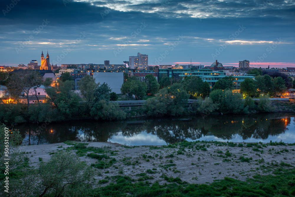 Magdeburg bei Nacht mit dem Fluss Alte Elbe im Vorderrung, beleuchteten Häusern im Hintergrund, der Johanniskirche im Abendlicht und Wolken und grünen Bäumen im Sommer