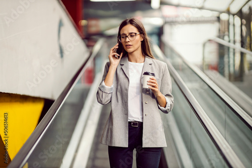 Canvas A busy young businesswoman dressed elegantly descending the escalator while holding takeaway coffee and talking on the phone with a client