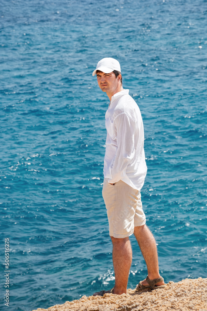 Back view of a man on a background of yellow rocky beach and blue sea. A guy in a white linen shirt, shorts, sandals and a cap. Travel vacation. Hands in pockets and looking away. Copy space.
