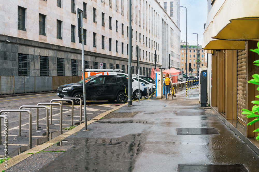 A girl in jeans with a yellow backpack on her back walks along the sidewalk in the rain on a yellow bicycle past a black electric car charging with a cable from an outdoor charging station in Milan