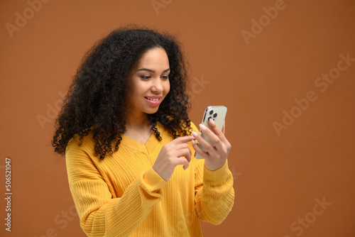 Attractive young multiracial woman web browsing on smartphone, millennial female chatting on social networks, using mobile app, texting online with friends, sharing news, isolated on brown