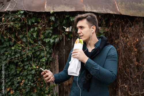 One man standing outdoor in autumn spring or winter day holding sandwich tourist wearing shirt eating while wait outdoor in front of old wall use mobile phone real people copy space fast food concept