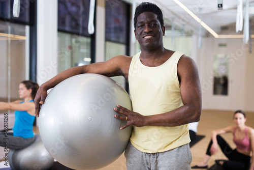 Smiling African American pilates instructor standing in fitness studio with gym ball during group workout..