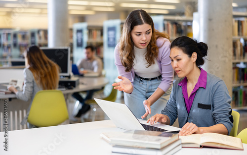 Two female friends work together on laptop and read books in a public library