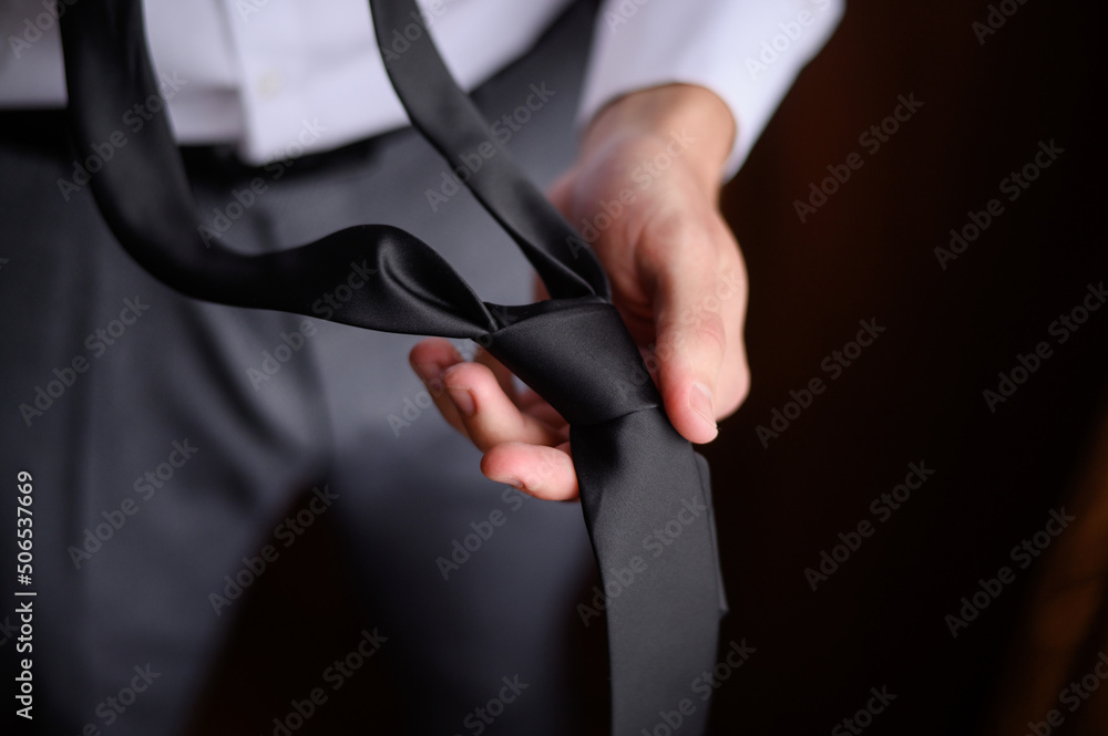 A man in a white shirt holds and adjusts his tie in his hands.Groom morning.