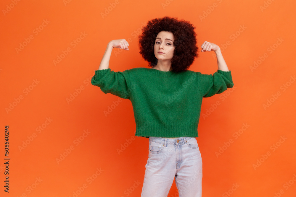 Strong confident woman with Afro hairstyle wearing green casual style sweater raising arms showing biceps, female strength and feminism. Indoor studio shot isolated on orange background. Strong confident woman with Afro hairstyle wearing green casual style sweater raising arms showing biceps, female strength and feminism. Indoor studio shot isolated on orange background.