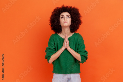 Hopeful woman with Afro hairstyle wearing green sweater holding hands in prayer and pleading, looking up with eyes full of hope, asking apology. Indoor studio shot isolated on orange background.