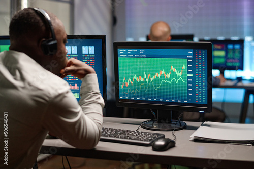 Back view of tired African American brokerage specialist sitting in front of computer monitors watching stock trading stats in office in evening