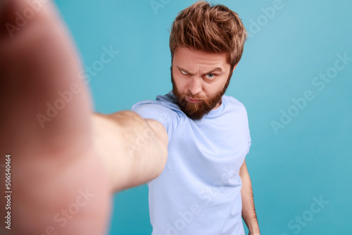 Portrait of handsome bearded man blogger making selfie or broadcasting livestream, looking at camera with angry aggressive expression, funny face. Indoor studio shot isolated on blue background.