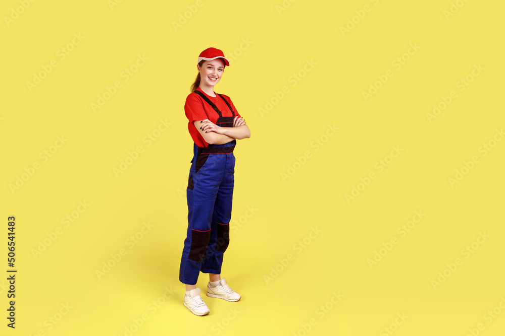 Full length portrait of happy smiling handy woman standing with crossed hands, having positive confident expression, wearing overalls and red cap. Indoor studio shot isolated on yellow background. Full length portrait of happy smiling handy woman standing with crossed hands, having positive confident expression, wearing overalls and red cap. Indoor studio shot isolated on yellow background.