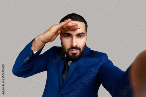 Positive handsome bearded businessman looking into distance with calm facial expression, point of view, wearing official style suit. Indoor studio shot isolated on gray background.