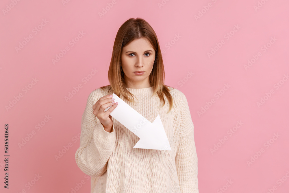 Portrait of dissatisfied unhappy blond woman holding down value fall arrow, looking at camera with sorrow and sadness, wearing white sweater. Indoor studio shot isolated on pink background. Portrait of dissatisfied unhappy blond woman holding down value fall arrow, looking at camera with sorrow and sadness, wearing white sweater. Indoor studio shot isolated on pink background.