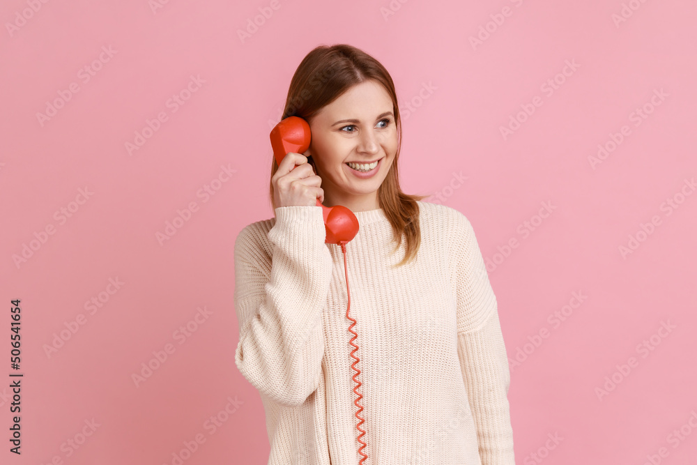 Portrait of happy satisfied blond woman talking on red retro telephone, having pleasant conversation, looking away with smile, wearing white sweater. Indoor studio shot isolated on pink background. Portrait of happy satisfied blond woman talking on red retro telephone, having pleasant conversation, looking away with smile, wearing white sweater. Indoor studio shot isolated on pink background.