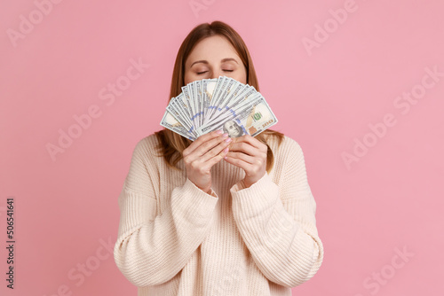 Tablou canvas Portrait of rich greedy blond woman holding big fan of dollars bills and smelling money with satisfied expression, wearing white sweater