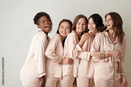 Positive young women in satin pajams laughing and hugging each other