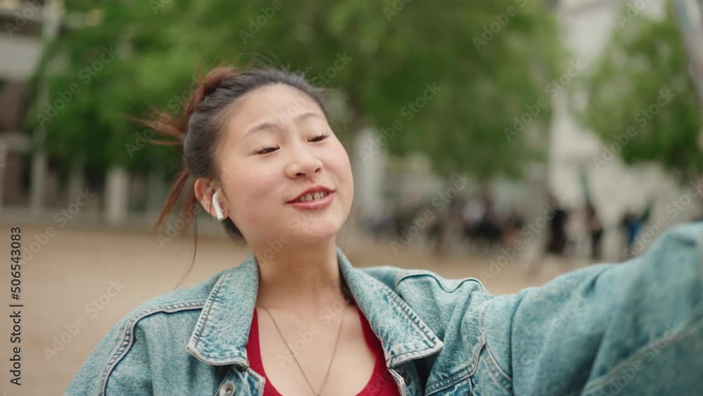 Attractive Asian girl wearing wireless earphones making video for her social media using smartphone outdoors. Female blogger creating content Attractive Asian girl wearing wireless earphones making video for her social media using smartphone outdoors. Female blogger creating content