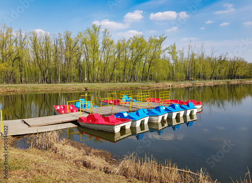 Colorful catamarans stand in a row at the wooden pier. Calm surface of the lake for water recreation. An empty embankment on an early spring or summer sunny morning